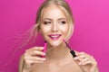 Blonde Model Girl holding Pink Lipstick Brush over Bright Magenta Background. Summer Beauty Woman Face Cosmetics Make up. Fashion Royalty Free Stock Photo