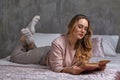 Blonde model in glasses, casual clothing. She is laying on bed and reading book in bedroom. Student, blogger. Interior