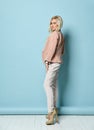 Model with long hair, in beige jacket, white overall, high heels. Smiling, standing against blue studio background. Full length Royalty Free Stock Photo