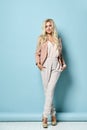 Model with long hair, in beige jacket, white overall, high heels. Smiling, standing against blue studio background. Full length Royalty Free Stock Photo