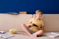 Blonde schoolboy boy plays video games, holds a gamepad, eats popcorn instead of learning lessons on blue background Royalty Free Stock Photo