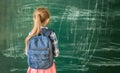 Blonde schoolgirl with backpack stands before chalkboard in classroom. Little girl with ponytail hairstyle prepares for