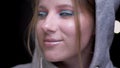 Blonde sportsgirl in hoodie with colorful make-up smiling shyly into camera on blurred lights background.