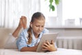 Blonde teenage girl lying on the couch plays a game on the tablet and communicates via video link with friends. Digital Royalty Free Stock Photo