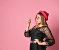 Teenage girl in sunglasses, black dress and red hat. She pointing at something by forefinger, posing on pink background. Close up Royalty Free Stock Photo
