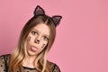 Blonde teenager in black dress, headband like cat ears, face painting. She showing her tongue, posing on pink background. Close up