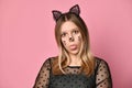 Blonde teenager in black dress, headband like cat ears, face painting. She showing her tongue, posing on pink background. Close up