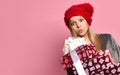 Blonde teenager in gray pajamas, red hat. She holding colorful gift boxes, kissing you, posing on pink background. Close up Royalty Free Stock Photo