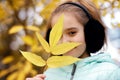 A Blonde Walks In An Autumn Park In Warm Headphones And A Blue Insulated Jacket, Covers Her Face With A Twig With Yellow Leaves