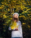 Blonde in white shirt and hat walks in beautiful park on sunny autumn day. Young pretty woman holds a branch with yellow leaves in Royalty Free Stock Photo