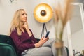 Blonde white woman wearing maroon shirt in living room putting pinger to chin thinking and looking up holding smartphone Royalty Free Stock Photo