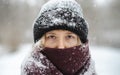 Blonde woman in beanie hat and nose covered in scarf looks at camera in winter snowfall. Nordic winter seasonal portrait, tranquil
