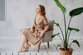 A blonde woman in a beige dress poses on a gray chair in a photo studio.