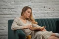 Blonde woman blogger in beige knitted dress and socks is sitting on gray sofa and reading book against white brick wall Royalty Free Stock Photo