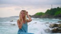 Blonde woman in blue dress enjoys serene sea breeze at rocky coast. Elegant lady hair, contemplates ocean waves