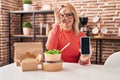 Blonde woman eating take away food showing smartphone screen smiling happy doing ok sign with hand on eye looking through fingers Royalty Free Stock Photo