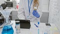 Blonde woman examines specimen in laboratory with microscope, flasks, and tablet Royalty Free Stock Photo