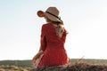 Blonde woman in hat and red dress is sitting on a haystack. View from the back