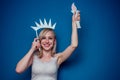 Blonde woman holding American flag with paper crown and torch Statue of liberty on a blue background in the studio .4th Royalty Free Stock Photo