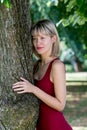 Blonde woman hugging a tree in park. Young girl in a red dress resting in nature, leaned against a tree. Royalty Free Stock Photo