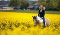 Laughing blonde woman sits on her horse in a blooming rapeseed field, houses and forest are out of focus in the background. Royalty Free Stock Photo