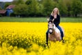 Laughing blonde woman sits on her horse in a blooming rapeseed field, houses and forest are out of focus in the background. Royalty Free Stock Photo