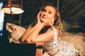 Blonde woman looks up dreamily sitting under floor-lamp in dark room. Vintage photo shows aristocratic girl in the interior of her