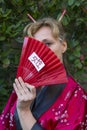 Blonde woman in oriental clothing covers her face with a red hand fan on which the word woman is written in Japanese Royalty Free Stock Photo