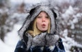 Blonde woman in plaid coat, fur hat and mittens in winter park sending air kiss. Portrait of beautiful girl in warm