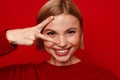 Blonde woman showing v-sign peace gesture near eye and smiling isolated over red wall Royalty Free Stock Photo