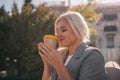 A blonde woman sits on a bench drinking coffee from a yellow cup. She is wearing a gray jacket and has her hair in a Royalty Free Stock Photo