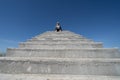 Blonde woman sits on top of concrete stairs platform against a blue sky. Negative space composition, with lots of copyspace for