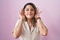 Blonde woman standing over pink background trying to hear both hands on ear gesture, curious for gossip