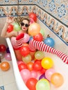 Blonde woman with sunglasses playing in her bath tube with bright colored balloons. Sensual girl with white red striped stockings Royalty Free Stock Photo