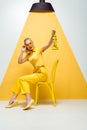 Blonde woman touching sunglasses while sitting on chair and holding vintage lamp on white and yellow Royalty Free Stock Photo