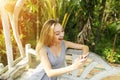 Blonde woman uses smartphone for chat on sunny day, background of sunshine green palms in Thailand, Phuket travel