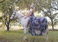 Blonde woman wearing blue floral dress seated on white horse in woods Royalty Free Stock Photo