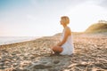 Blonde woman wearing long white dress sit at empty beach and look at the sea