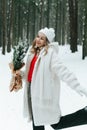 Blonde woman wears white fur coat and knitted hat posing with fir tree in winter forest. Winter fashion Royalty Free Stock Photo