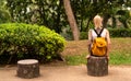 A blonde with a yellow backpack sits in a green park. Wears a red bandage, arms with tattoos. Asia travel