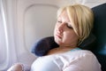 A blonde young adult woman uses a neck pillow to sleep and nap on a long airplane flight. Woman is in the window seat Royalty Free Stock Photo