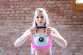 Blonde young muscular woman with sporty looks doing high pull exercise with kettlebells on hard training at the garage