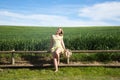 Blonde, young, pretty farm woman, sitting on a wooden fence. In the background a green wheat field and blue sky. The woman is