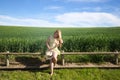 Blonde, young, pretty farm woman, sitting on a wooden fence. In the background a green wheat field and blue sky. The woman is