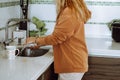 Blonde young woman in home clothes wash mugs in sink under running water after eating and tidy up modern kitchen side Royalty Free Stock Photo