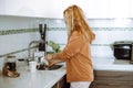 Blonde young woman in home clothes wash mugs in sink under running water after eating and tidy up modern kitchen side Royalty Free Stock Photo