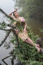 Blooming gorgeous lady in dress of flowers lying on tree over water in the morning fog