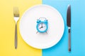 Blue alarm clock, fork, knife and empty plate on colored paper background. Intermittent fasting concept