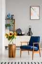 Blue armchair next to wooden table with sunflowers in grey flat interior with poster.