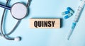 On a blue background, a stethoscope, a syringe and pills and a wooden block with the word QUINSY. Medical concept